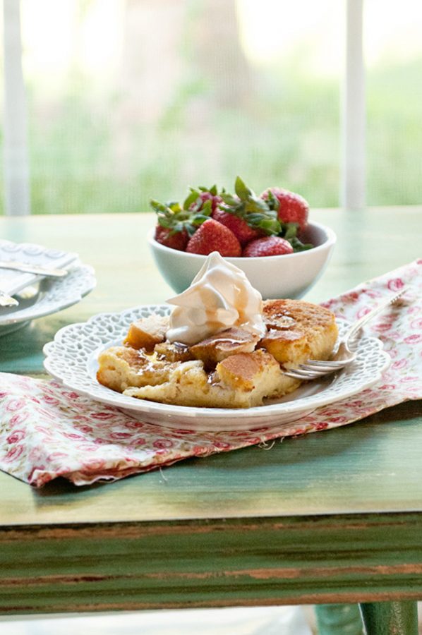 French Toast Casserole on a white plate by a window. A bowl of strawberries and cloth napkin.