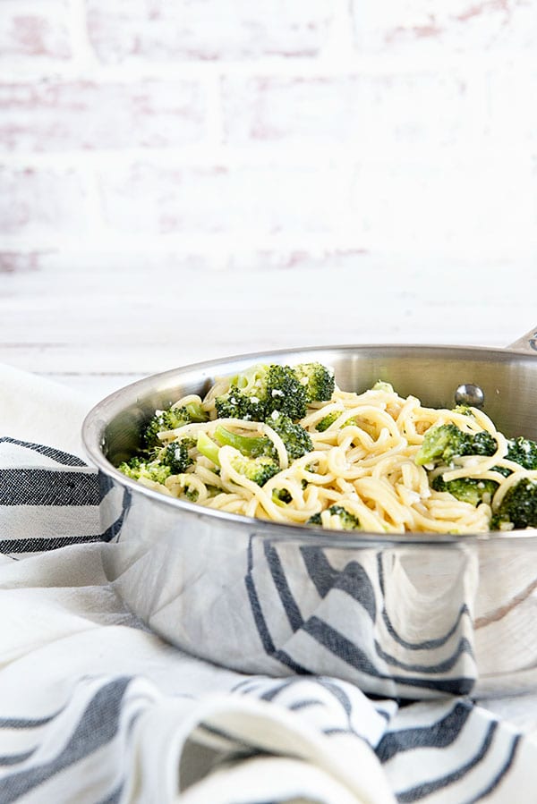 Spaghetti and broccoli in a silver pan with a white brick background