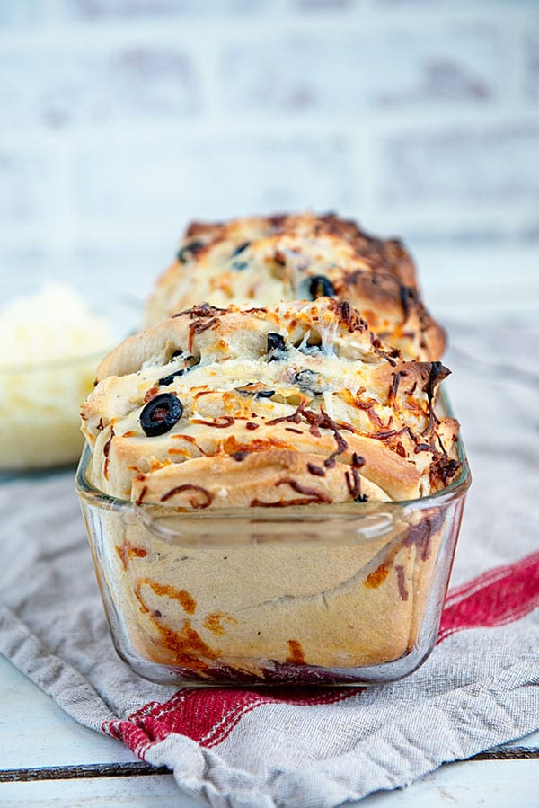 A cheese, bacon and olive pull apart bread in a glass baking dish with a glass bowl of shredded cheese