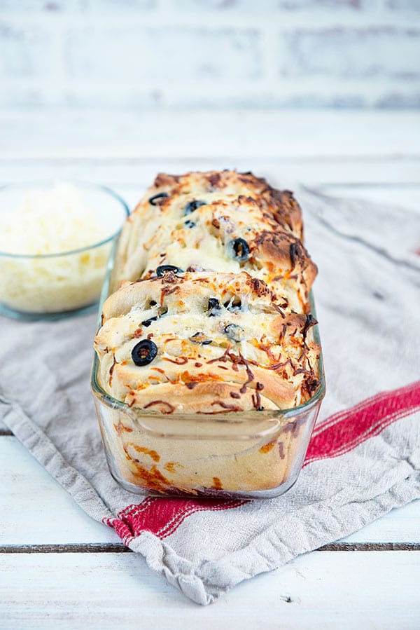 a loaf of cheesy bread in a glass pan with a bowl of shredded cheese