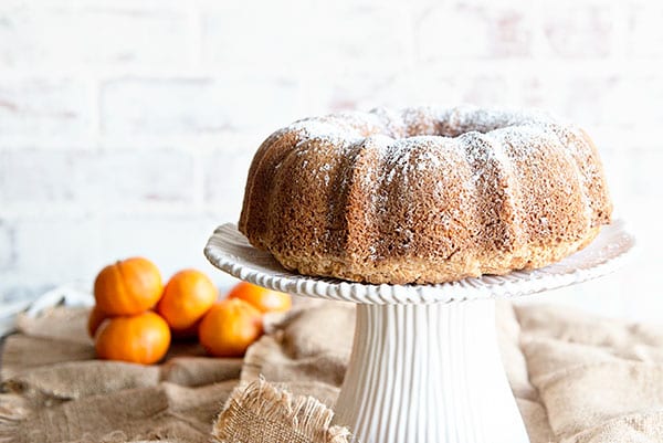 White background with an orange juice bundt cake on a white cake stand. Oranges are in the background on a burlap cloth.