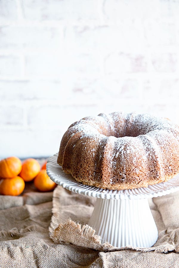 White faded brick background with a bundt cake on a white cake stand. Clementines are in the background on burlap.