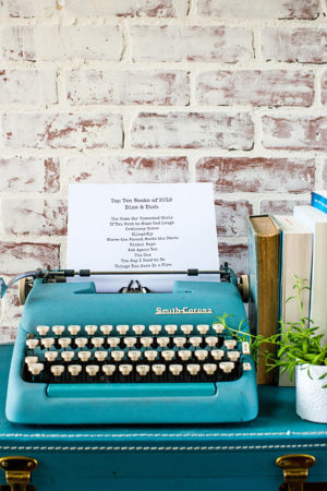 Typewriter sitting on a suitcase with books and a plant nearby and a list of favorite books read in 2019