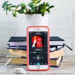 iPhone screen with Mercy House Audiobook on it, a stack of books and a green leaf plant