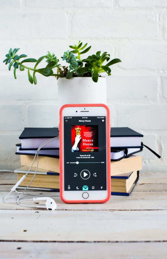 iPhone screen with Mercy House Audiobook on it, a stack of books and a green leaf plant