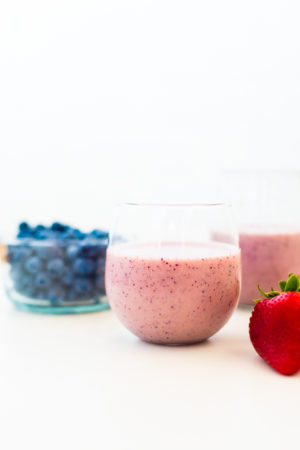 white background with two glasses filled with berry smoothies and a bowl of blueberries